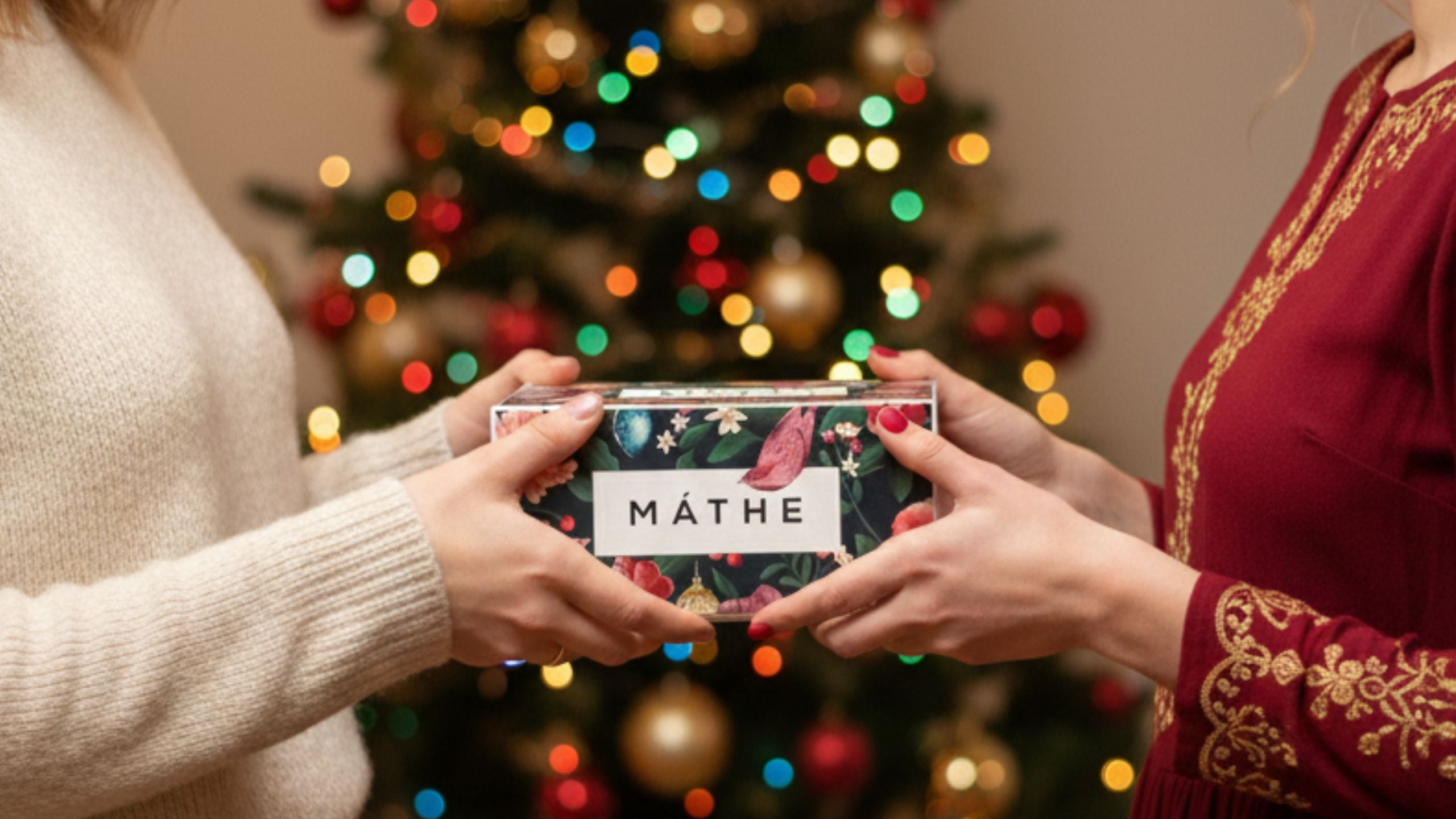 Manos entregando un regalo de Máthe en caja navideña frente a un árbol decorado, simbolizando regalos personalizados y cosmética natural hecha a mano